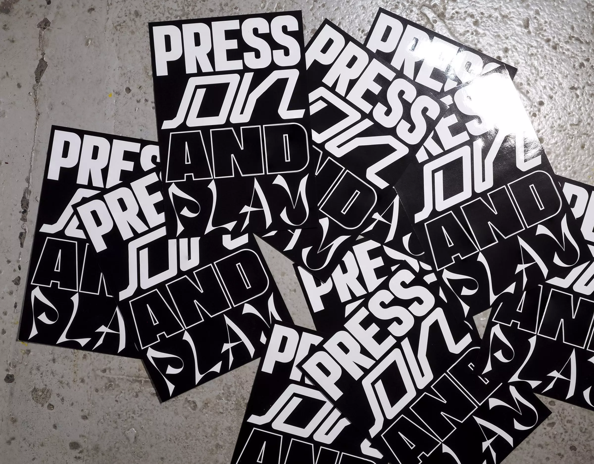 Black flyers with white type which reads 'Press On And Play', scattered on a grey concrete floor.
