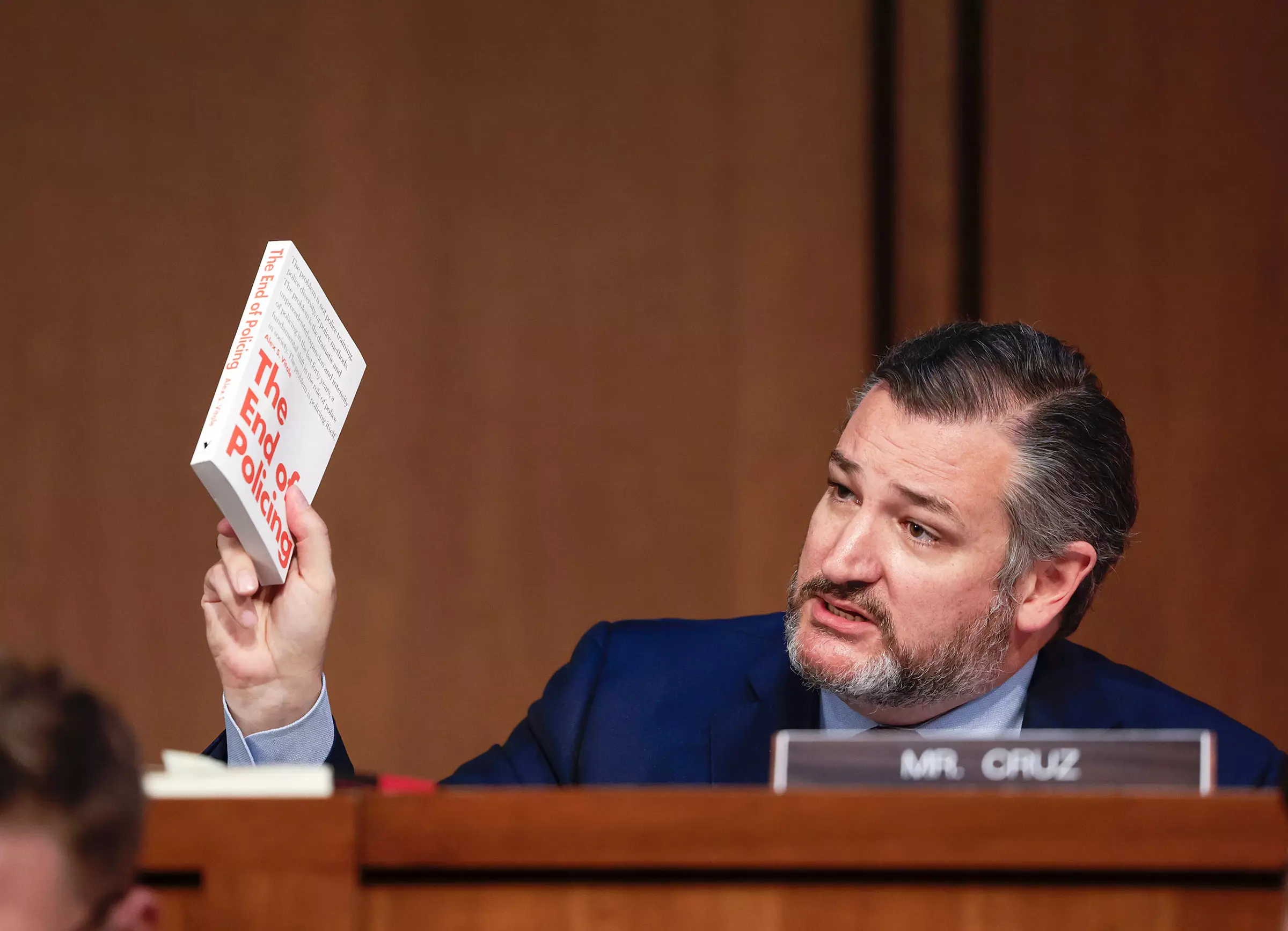 An image of Senator Ted Cruz sitting in the Senate Judiciary Committe for the the Supreme Court confirmation hearing of Ketanji Brown Jackson. Cruz sits to the right of the image at a wooden desk, facing the camera. With his right hand he holds up a book, with 'The End of Policing' visible in large orange text on its cover.